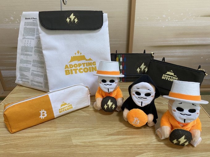 A display of "Adopting Bitcoin" merchandise including a white drawstring bag, pouches, and three plush dolls in orange and black outfits with mask faces.