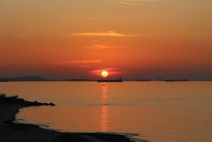A serene ocean sunset with an orange sky, anchored ships silhouetted on the horizon, and a sandy beach stretching along the foreground.