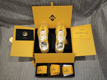 A yellow shoebox with a geometric logo opens to reveal gold and silver metallic sneakers, accompanied by a yellow pouch and additional small compartments containing accessories.
