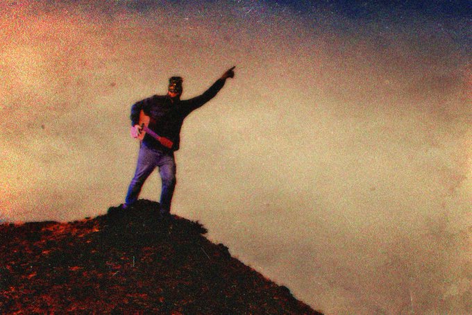 A vintage-style photo of a man with a guitar standing on a hilltop pointing toward the sky against a hazy sunset.