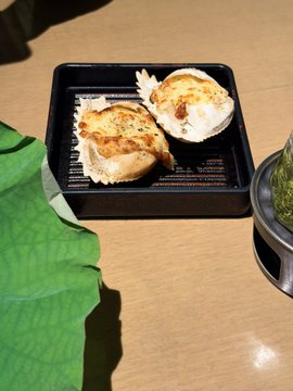 A close-up photograph of two pieces of grilled, seasoned food served in a black rectangular dish on a wooden table, with a green cloth and a bowl of green garnish nearby.