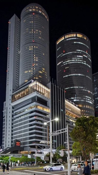 A nighttime cityscape photo of two large illuminated cylindrical skyscrapers (appears to be JR Central Towers in Nagoya) with cars and pedestrians on the street below.
