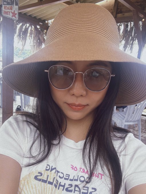 A selfie of a woman wearing a wide-brimmed tan sun hat and round sunglasses, with the text "SEASHELLS COLLECTING THE BEACH" (mirrored) visible on her white t-shirt.
