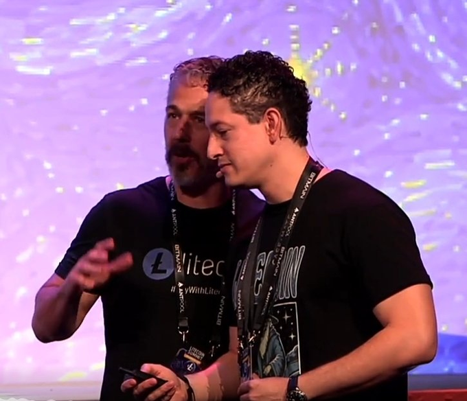 Two men stand on stage with lanyards, one wearing a black "Litecoin" t-shirt with the hashtag "#PayWithLitec[oin]" while the other sports a graphic tee featuring what appears to be Elon Musk; the post humorously plays on the idea of sensing insider knowledge about "Charlie," a suspected Litecoin intern, tied to a whimsical bit about intuition in his "plums."