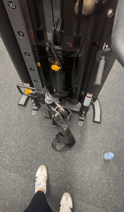 A first-person view of someone standing in white sneakers in front of a black cable weight machine at a gym, with weights from 7 to 10 visible and a small water bottle on the floor.