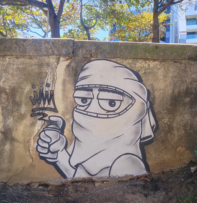 Black and white graffiti mural of a mummy character wearing a head covering and holding up a crown, painted on a concrete wall outdoors.