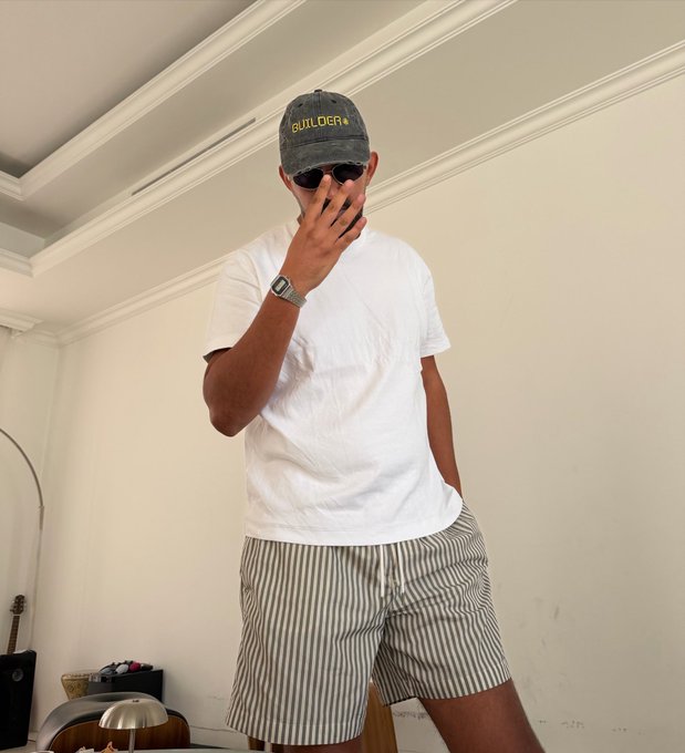 A man poses indoors wearing a "BUILDER" cap, sunglasses, a white tee, and striped shorts, his hand covering part of his face, with a guitar and lamp visible in the background—matching the post's reference to "Builder Ben Todar."