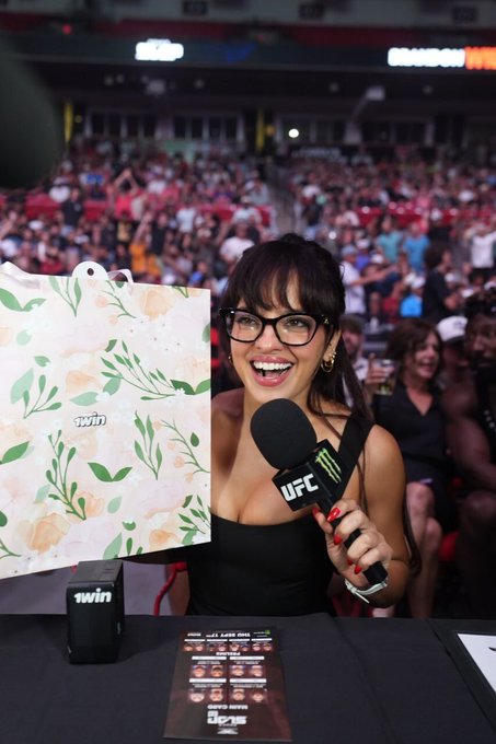 Nina Drama holds a floral gift bag branded with "1win" while smiling into a UFC microphone, surrounded by a lively arena crowd. A promotional flyer on the table highlights 1win's VIP program, tying the image directly to her live-streamed unboxing and endorsement of the brand.
