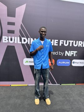 A young man in a blue shirt stands smiling at a "Building the Future" event backdrop sponsored by NFT.monks, AURUM, and Bitget Wallet, posing with a shaka hand gesture. The image visually represents the post about Sui Network users moving stablecoins for $0, likely capturing the speaker at a crypto/blockchain event promoting the network's zero-fee stablecoin transactions.