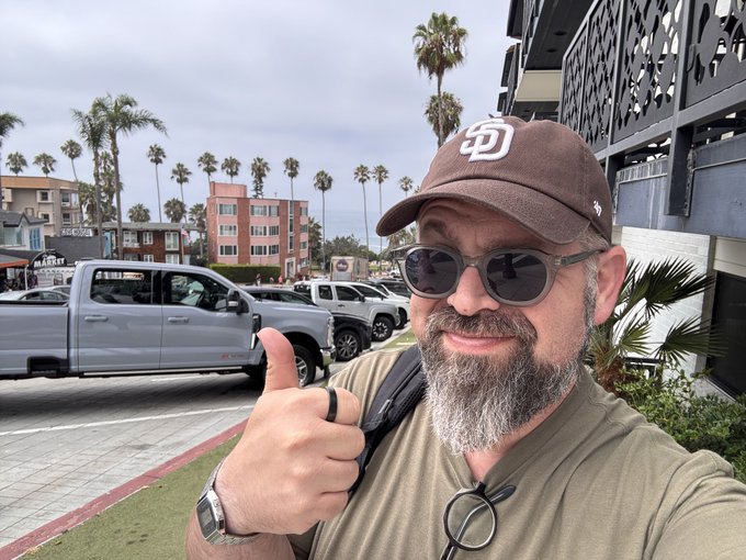 A bearded man wearing sunglasses and a brown "SD" Padres cap gives a thumbs-up in what appears to be Coronado, CA (America's nicest city), with palm trees, coastal buildings, and parked vehicles in the background—greeting the Arbitrum community with good morning vibes.