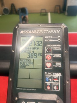 The digital display of an Assault Fitness machine shows '21:32 Results' with 'CALORIES 389.26' and 'STROKES 150320' in a gym setting.