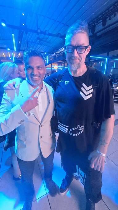 A photograph of two men smiling and posing together at what appears to be an event with blue lighting, with one wearing a white blazer and the other in a black graphic t-shirt and glasses.