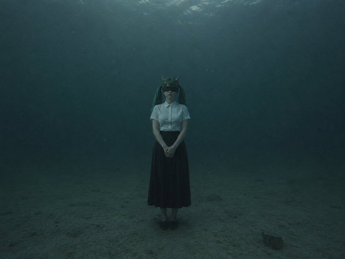 An artistic, surreal image shows a person in a school uniform standing underwater with small horns on their head and sunglasses over their eyes.