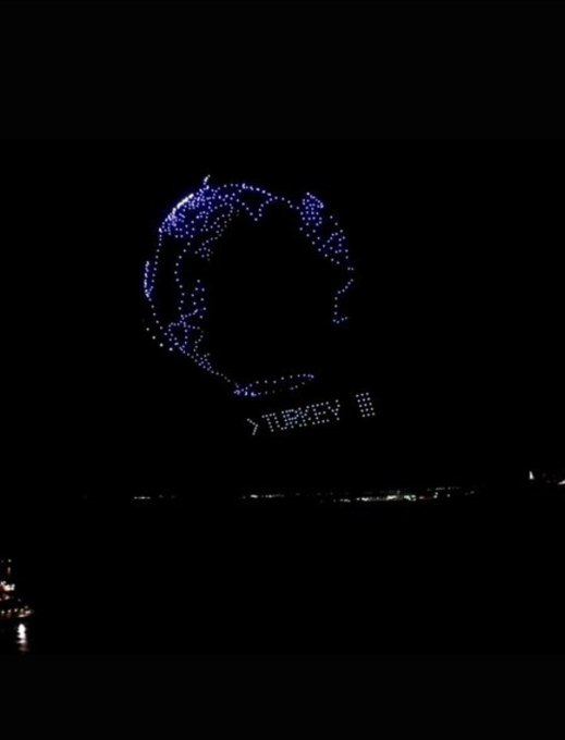 A nighttime drone light show displays a blue globe made of illuminated dots, symbolizing global community and inclusion, hovering above a cityscape with the partial text "TREK" visible on a building below.
