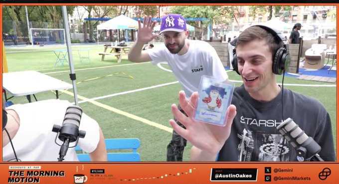 The image shows two men at an outdoor podcast setup labeled "THE MORNING MOTION," with one wearing a "STAFF" shirt and a NY Yankees cap waving, while another wearing headphones holds up a signed Pudgy Penguins "Kakawow" card featuring Luca Netz's autograph, fitting the post about Gemini showcasing the card live from Collector Park.