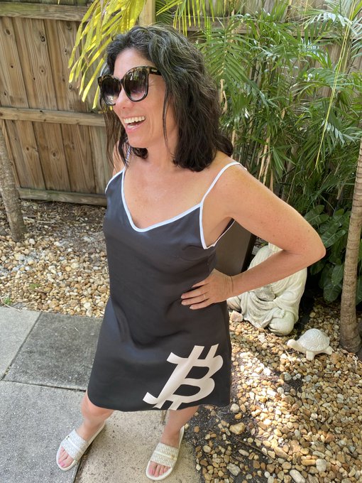 A smiling woman with sunglasses wears a black slip dress featuring the Bitcoin '₿' logo, posing in a garden area with plants and a wooden fence.