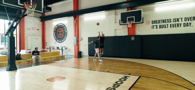 Two people play basketball on an indoor court while a person sits on a bench watching; the wall displays the text 'GREATNESS ISN'T OVER. IT'S BUILT EVERY DAY.'