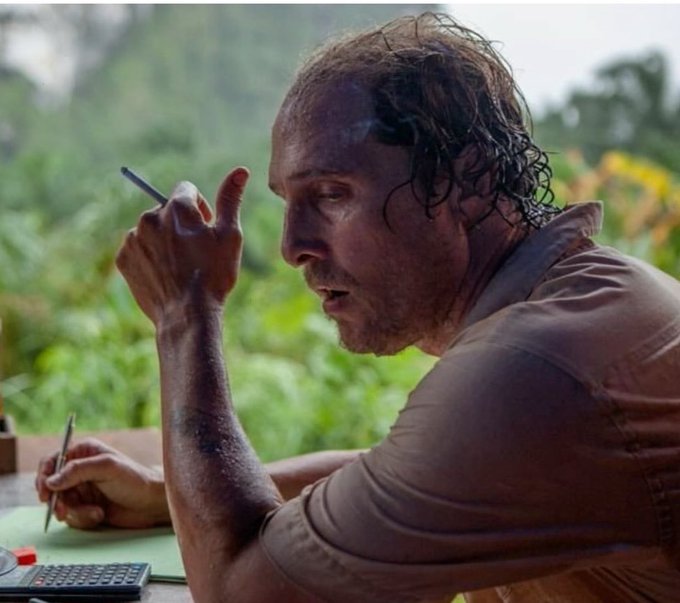 A cinematic shot of a sweaty, disheveled man in a tan shirt smoking a cigarette while holding a pen over paper, with a calculator visible on the table in a lush green outdoor setting.