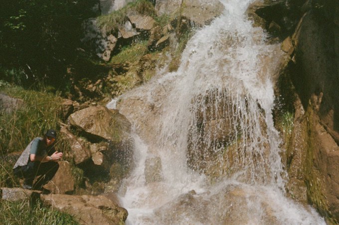 A person crouches on rocky terrain next to a rushing waterfall surrounded by green vegetation.