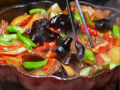 A close-up of a simmering stir-fry or stew containing wood ear mushrooms, green peppers, and red peppers being stirred with chopsticks.