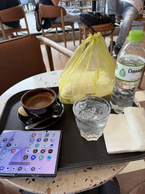 A photograph of a café table with a tray holding a cup of espresso, a glass of ice water, a yellow takeout bag, a Dasani water bottle, napkins, and a foldable smartphone displaying its home screen apps.