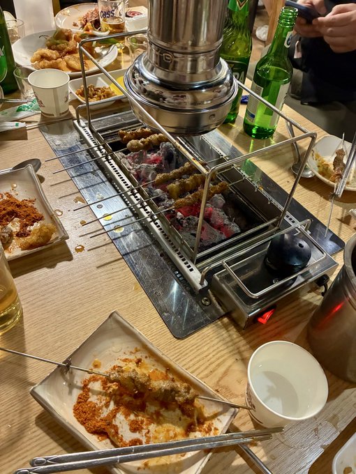 A table at a Korean BBQ restaurant with a charcoal grill, plates of food, green soju bottles, and a partially eaten plate of fried chicken with red seasoning.