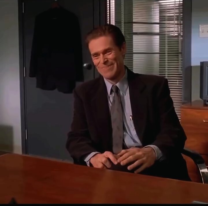 The image features a man sitting at a desk, wearing a suit and tie, and smiling as he looks directly at the camera. The background includes a window that allows natural light into the room, suggesting an office setting. There are also two TVs in the scene - one on the left side of the frame and another on the right side. The man's attire and demeanor give off a professional vibe, which could be related to the post about making money or discussing business matters.