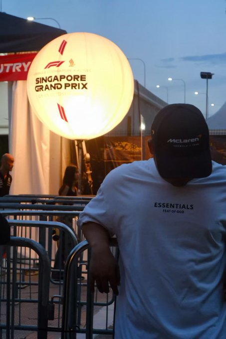 A person in a white 'ESSENTIALS FEAR OF GOD' t-shirt and McLaren cap leaning on metal barriers, with a glowing 'SINGAPORE GRAND PRIX' lantern and a 'COUNTRY' sign visible in the background at dusk.