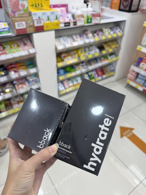 A first-person view of a hand holding two black skincare product boxes labeled 'b.back hydrate recovery' inside what appears to be a pharmacy or convenience store with various items visible on the background shelves.
