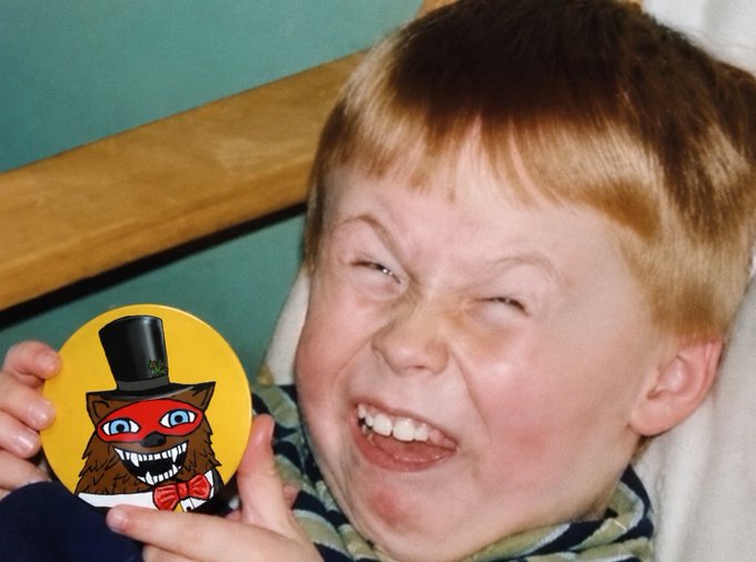 A young red-haired boy grins widely while holding up a round yellow sticker featuring a cartoon cat with a top hat and red mask.