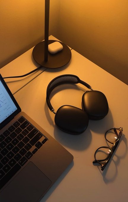 A still life of a workspace featuring dark over-ear headphones resting on a light desk beside a laptop, reading glasses, and a white earbuds case near a lamp base, setting a calm, quiet Sunday morning mood.