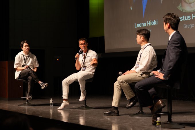 Four panelists sit on stools on a dimly lit stage at the WebX Asia conference in Tokyo, with one speaker addressing the audience using a microphone while a large screen behind displays names including "Leona Hioki" and partial text reading "stus" and "Grego," reflecting the event's buzz around institutional adoption and the convergence of TradFi with blockchain-based businesses.