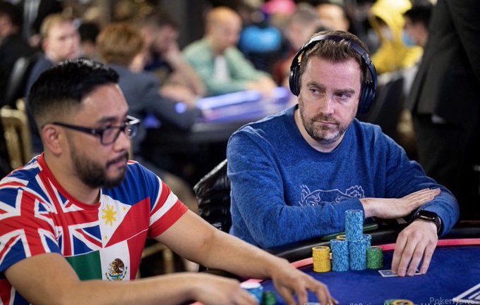 Two men seated at a poker table with chips stacked in front of them; one wears a Union Jack/Mexican flag shirt and the other wears a blue sweater and headphones.