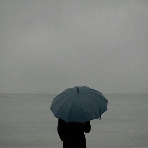 A moody, minimalist photograph of a silhouette holding a gray umbrella against a foggy, overcast seascape.