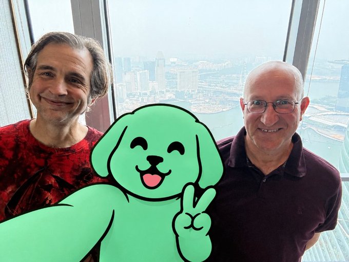 Two men pose for a photo with a green cartoon dog emoji sticker holding a peace sign, with a city skyline visible through the window behind them.