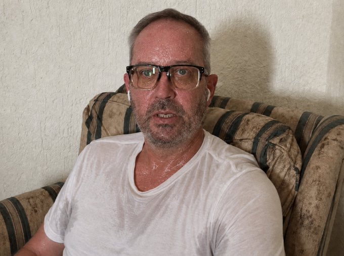 A middle-aged man with glasses, a beard, and a sweat-soaked white t-shirt sitting on a striped couch, appearing visibly exhausted.