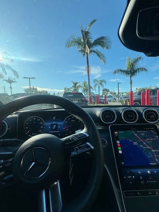View from behind the wheel of a Mercedes-Benz, with palm trees visible through the windshield and a navigation map showing Los Angeles/Long Beach displayed on the central infotainment screen—a perfect "Good Morning Web3" vibes shot, though the luxury ride suggests the poster's $100 breakfast might pair nicely with the upscale commute.