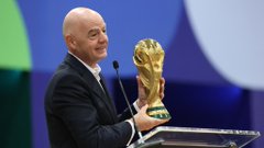 Gianni Infantino, wearing a dark suit, smiles while holding up the FIFA World Cup trophy at an event.