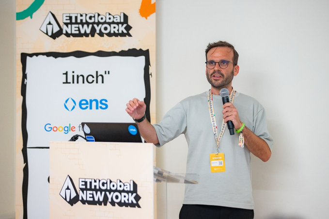 The image features a man standing at a podium, giving a speech or presentation. He is wearing a gray shirt and glasses, and he has his hands raised while speaking into a microphone. The backdrop of the scene includes a sign that says "ethGlobal New York" and another sign with the word "1 inch". There are also two laptops on display in front of the man, one near him and the other further away from him.