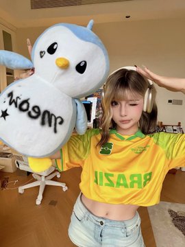 A young woman wearing a yellow Brazil jersey and gold headphones holds up a large Pudgy Penguins plush toy that reads "Woonmi" upside down, posing with her arms raised in a playful celebration pose. The image perfectly matches the post's game-day checklist, showing the fan geared up with her Brazil shirt and cute penguin mascot for what appears to be a World Cup match.