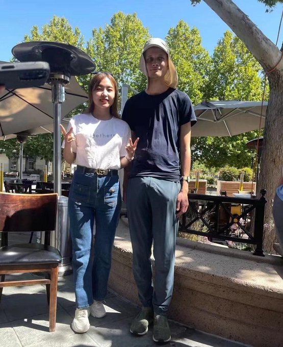 Two young people pose for a photo outdoors at what appears to be a cafe patio, with one making a peace sign while wearing a white 'tether' t-shirt and jeans.