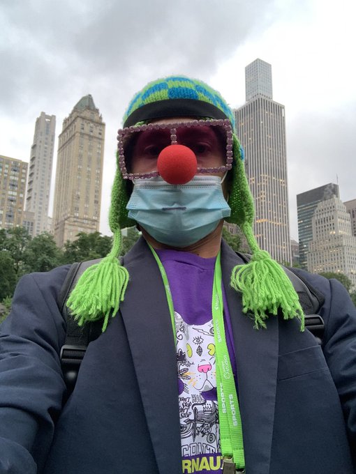 A person in a selfie wears a clown disguise with a red nose, novelty glasses, surgical mask, and green tassel hat, standing before a city skyline.