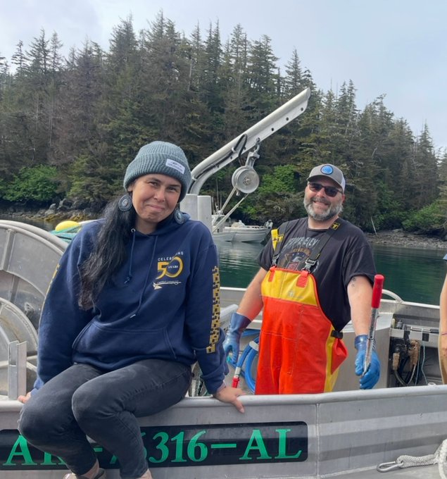 Two people, a woman in a blue 'Kellogg's 50' hoodie and beanie and a man in an orange waterproof bib and gray cap, smiling on a small boat in a wooded coastal area with Alaska registration 'AK 7316-AL' visible.