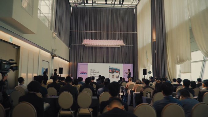 An audience attends a presentation in a modern event space with high ceilings, where a screen displays "Being Embraced by the Light."