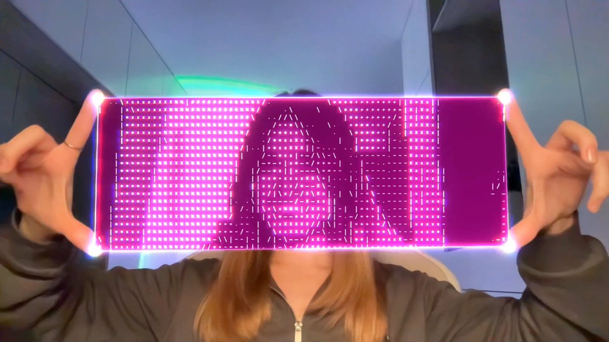 A person holding up a transparent LED display panel in front of their face, with the screen showing purple and pink pixelated patterns and text in front of a blue-lit background.