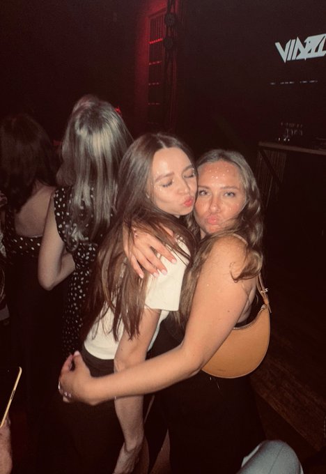 Two young women pose together cheek-to-cheek in a dimly lit nightclub setting, with a 'VINEZ' sign visible in the background.