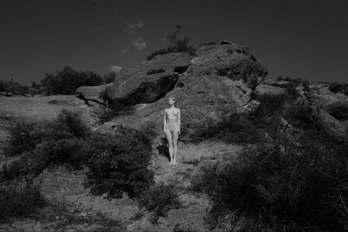 A black and white photograph depicts a nude woman standing amidst a rocky, desert landscape, which serves as a visual metaphor for the "Strength of the Roots" mentioned in the post.