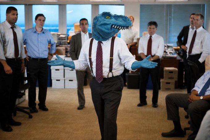 A person dressed in a business shirt, tie, and suspenders, with a cartoonish blue dinosaur head and matching blue-gloved hands, stands with arms outstretched in an office setting surrounded by several suited colleagues.

The image humorously visualizes the "raptor" or "shark-tank-like" salesman energy referenced in the post, playing on the stock ticker context as the dinosaur-costumed figure pitches to onlookers in a corporate office.