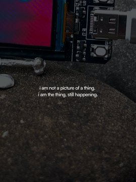 A small electronic device with a screen displaying pink and blue hues rests on a stone-like surface, accompanied by the caption "i am not a picture of a thing, i am the thing, still happening." This contemplative text mirrors the post's anticipation for the upcoming collaboration with @onchainac, suggesting the forthcoming work is a living, evolving piece rather than just a static image.