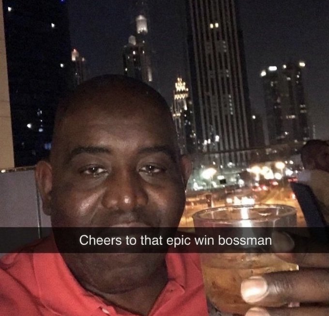 A selfie of a smiling bald Black man in a red shirt holding a glass of liquor, with the caption "Cheers to that epic win bossman" and illuminated city skyscrapers at night in the background.